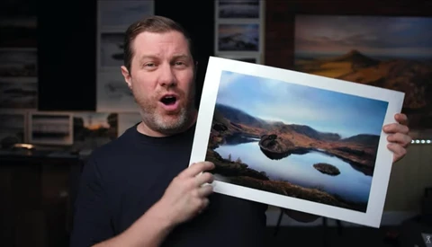 Man holding a framed landscape photograph of a mountain lake with reflective water.