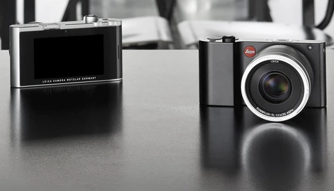 Two Leica digital cameras displayed on a dark surface, shown in profile view with their distinctive designs highlighted.