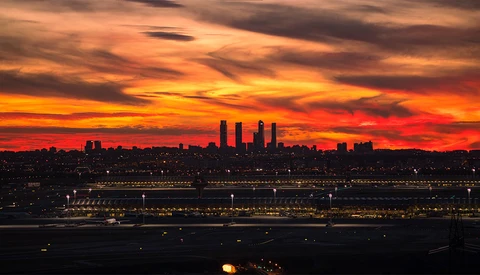 Dramatic sunset over a waterfront city skyline with orange and red clouds illuminating illuminated buildings and a large structure below.