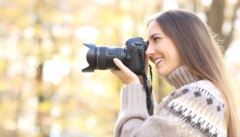 Woman with long brown hair holding a DSLR camera up to her face, smiling while framing a shot outdoors.
