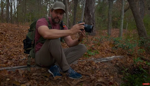 Photographer in hiking gear crouching in a forest while holding a compact camera at eye level.