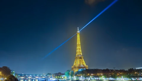 Eiffel Tower illuminated at night with a blue laser beam shooting upward into the dark sky, reflected in the Seine River below.