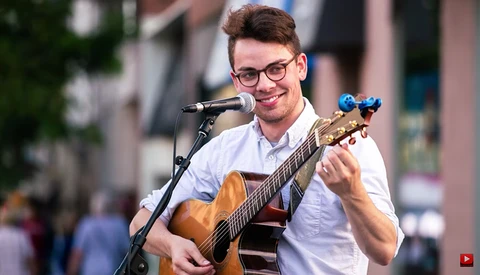 Musician performing outdoors with acoustic guitar and microphone, wearing glasses and light blue shirt.