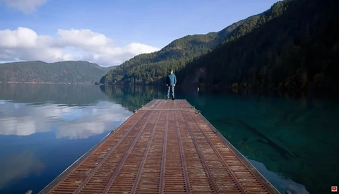 Person standing at the end of a wooden pier extending into a calm alpine lake surrounded by forested mountains.