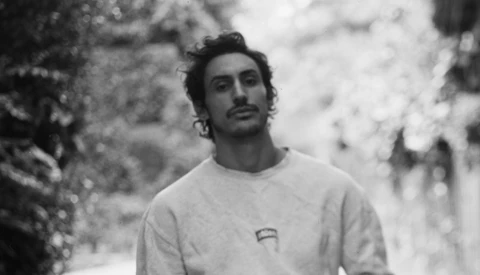 Man with long dark hair and mustache wearing light-colored crewneck shirt, photographed outdoors in black and white.