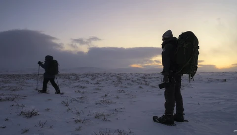 Three hikers with large backpacks trek across a snowy landscape during twilight.