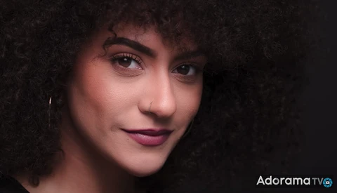 Portrait of a woman with curly dark hair against a dark background, lit with warm side lighting.