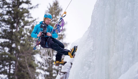 Ice climber ascending a frozen waterfall with crampons and ice axes, wearing blue jacket and helmet.