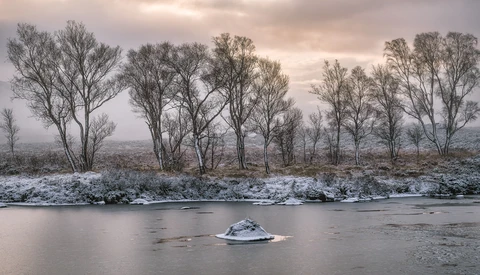 Bare trees line a snowy riverbank with a small ice-covered island in the foreground and misty hills beyond.