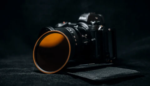 Camera lens with orange rim positioned in front of a DSLR body against a dark textured surface.