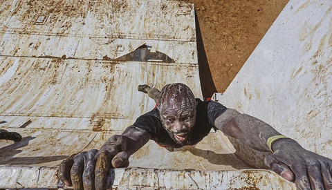 Mud-covered person climbing over a white wooden obstacle wall during an outdoor endurance event.