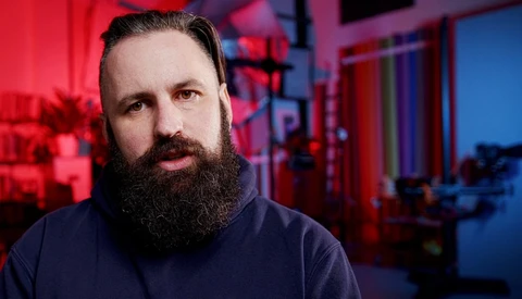 Bearded man with dark hair in a dark hoodie, photographed against red and blue neon lighting.