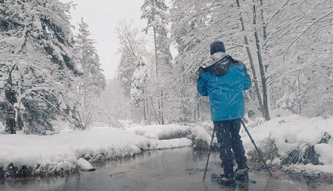 Cross-country skier in blue jacket gliding on snow-covered frozen stream surrounded by frost-laden evergreen forest.