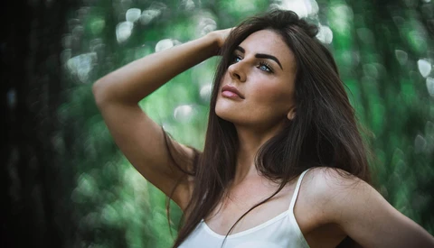 Woman with long dark hair gazing upward in a forest setting with blurred green background.