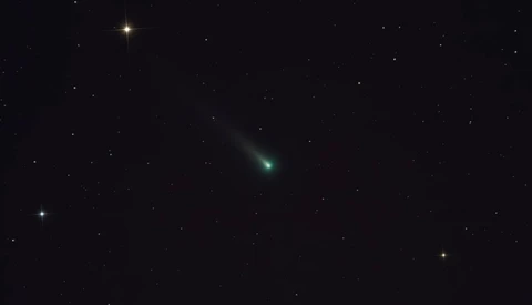 Comet with greenish coma and tail streaking across dark starfield at night.