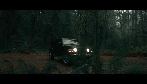 Black Jeep with headlights on driving through a dark forest at dusk or night.