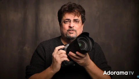 Man holding a camera lens toward the viewer against a neutral backdrop.