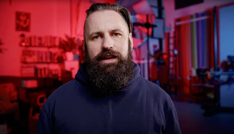Man with full beard wearing dark hoodie against red neon-lit background.