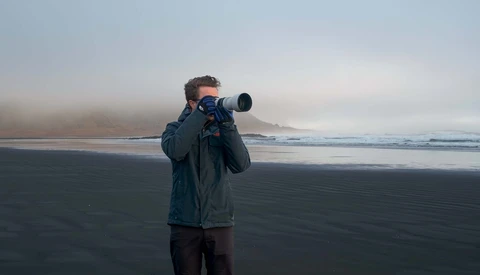 Photographer holding a telephoto lens on a misty beach with dark sand and calm water.