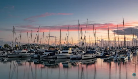 Sailboats with tall masts moored in calm water at sunset, their reflections mirrored in the still harbor.