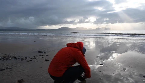 Person in red jacket crouching on wet sand at beach with mountains in background under dramatic cloudy sky.