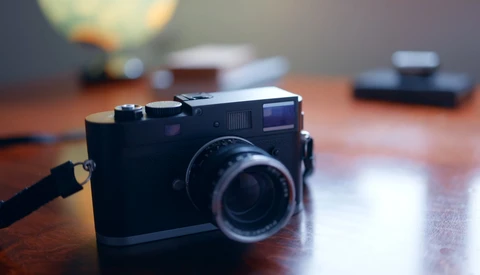 Black rangefinder camera with leather strap on wooden desk with blurred photography equipment in background.