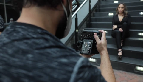 Photographer holding a mirrorless camera with articulating screen, capturing a portrait of a woman posed on illuminated stairs.