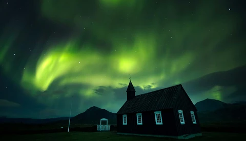 Red wooden church silhouetted against vibrant green aurora borealis dancing across night sky.