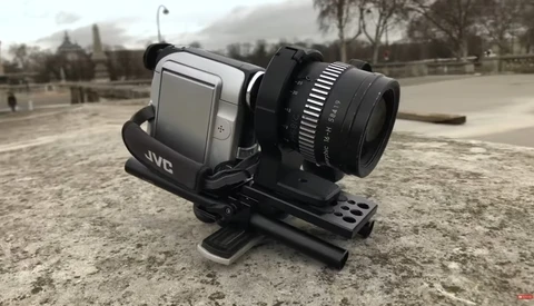 JVC compact camcorder mounted on a stabilizing rig with external lens attachment, positioned on ground-level support.