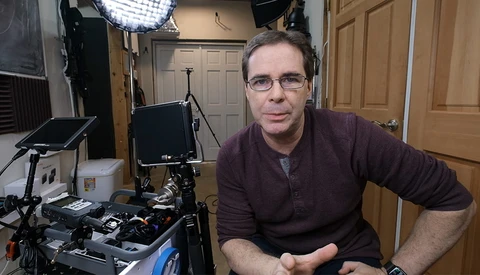 Man in glasses and burgundy cardigan seated in a professional video production studio setup.