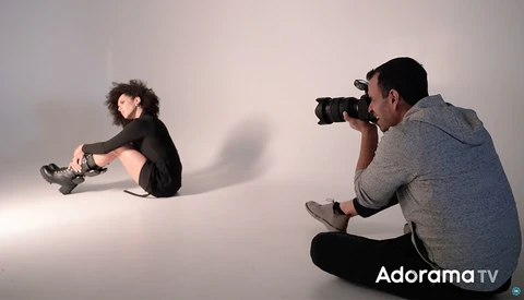 Photographer shooting a portrait of a seated subject against a white backdrop with dramatic side lighting.