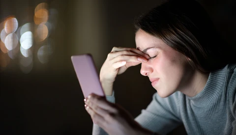 Woman viewing smartphone screen at night with hand to face, warm ambient light in background.