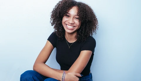 Woman with curly dark hair smiling at camera while seated against light blue background.