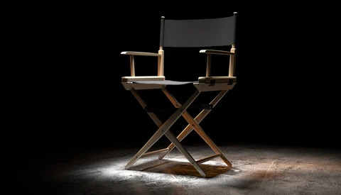 Director's chair with black canvas seat and backrest on wooden X-frame, spotlit against dark background.