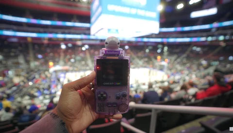 Hand holding a purple Game Boy Advance at a crowded indoor arena with illuminated scoreboard overhead.