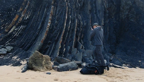 Photographer standing on beach with camera, framing dramatic columnar basalt rock formation.