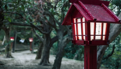 Dark red wooden lantern-style structure with illuminated windows hanging from a post among leafy trees.