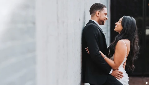 Couple in formal attire leaning against a white brick wall, looking at each other intimately.