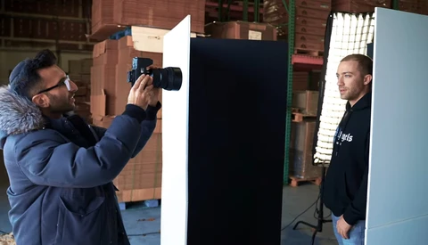 Photographer shooting a portrait setup with a large black reflector and model in an industrial studio space.