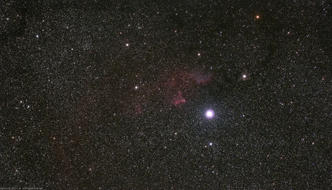 Deep space photograph showing a bright star surrounded by nebulosity and countless smaller stars against a dark starfield.