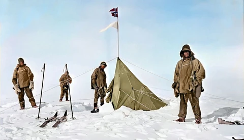 Five people in tan cold-weather gear standing around a canvas tent with a flag on a snowy landscape.