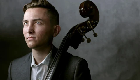 Cellist in formal attire holding an instrument against a neutral gray background.