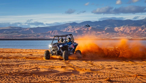 Off-road vehicle kicking up orange dust while driving across sandy desert terrain near a blue lake with mountains in the background.