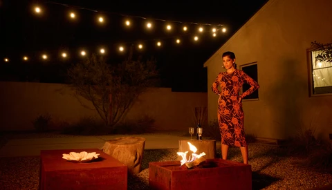 Woman in red patterned dress standing in lit courtyard with fire bowls at night.