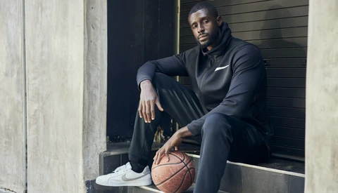 Male athlete in black athletic wear seated with basketball in an urban corridor.
