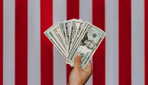 Hand holding a fan of U.S. dollar bills against a red and white striped background.