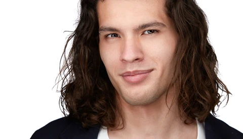 Man with long dark wavy hair wearing a dark blazer over a white shirt against a white background.