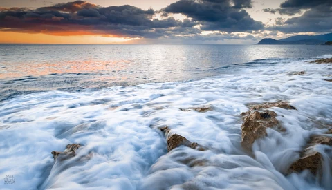 Waves crash over rocky beach at sunset with dramatic orange and blue clouds reflected in the water.