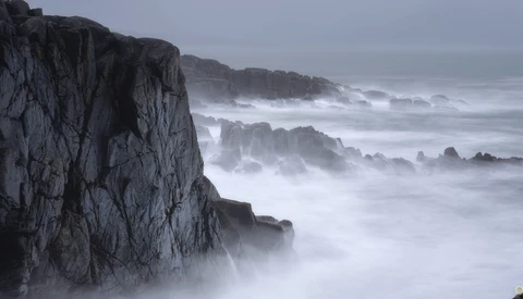 Dramatic rocky coastline with crashing waves and mist shrouding distant cliffs.