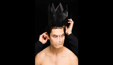Shirtless man adjusting a black crown-shaped headpiece against a black background.